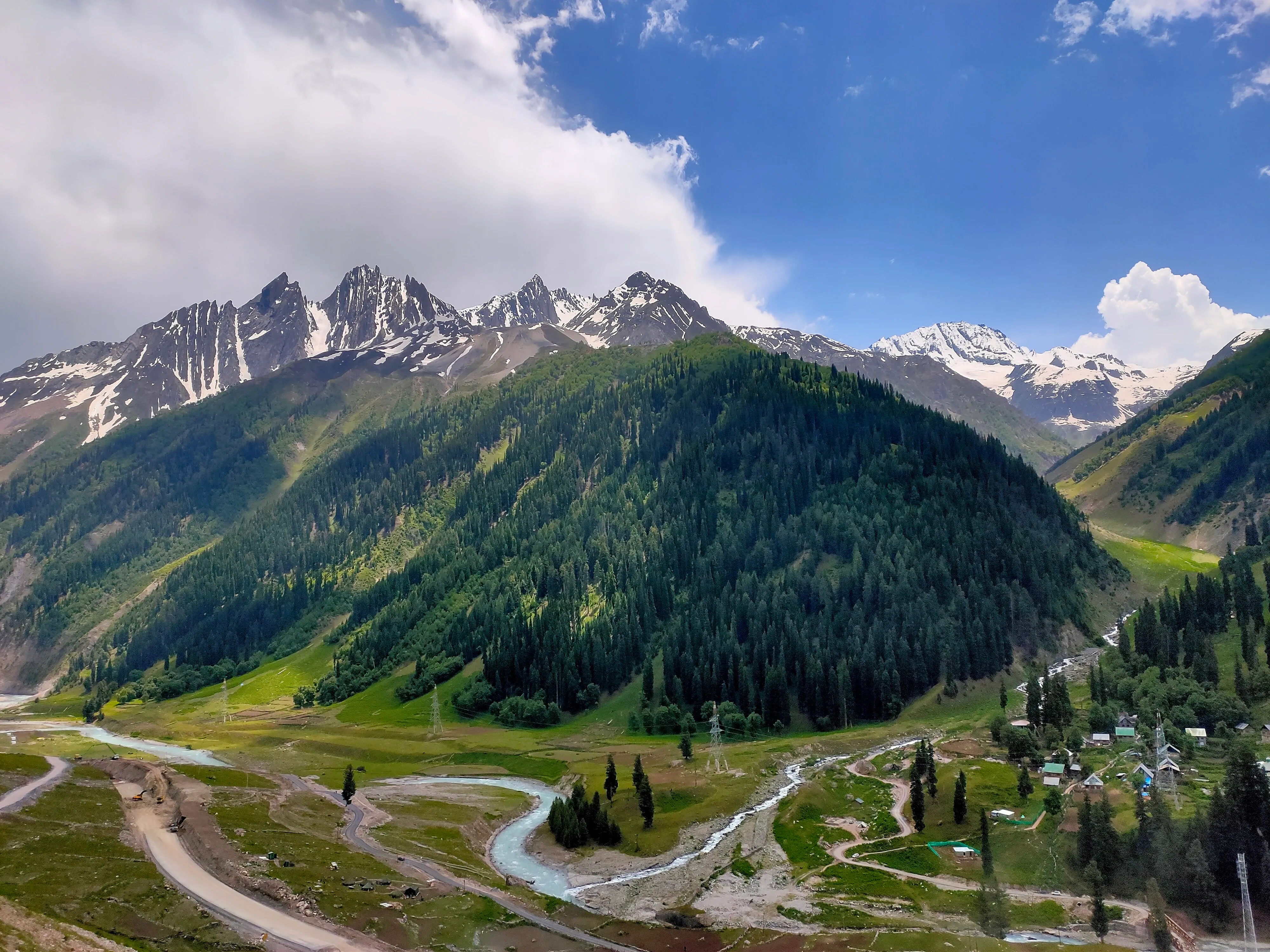 A green valley with pine forests, a river, and snow-capped peaks
